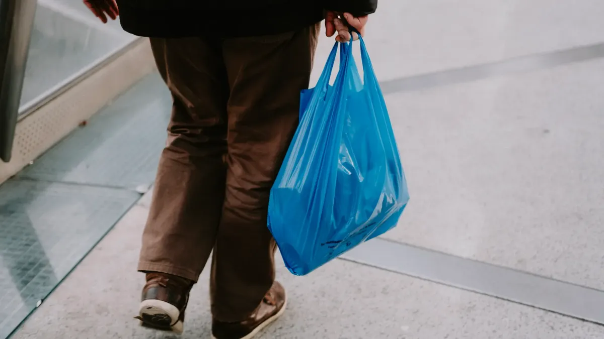 Rear view of a man carrying groceries in a blue plastic bag on an urban sidewalk.